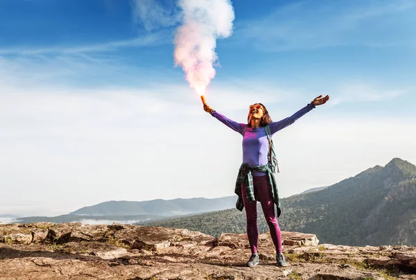 A woman in the wild mountains gives a distress signal SOS using Falsch feuer torch from which comes a bright flame and orange smoke, Concept of emergency situation during hike in the woods