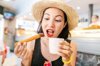 Happy Cheerful girl in hat eating traditional spanish delicious churros, a fried pastry with chocolate in cafe in spain clipart