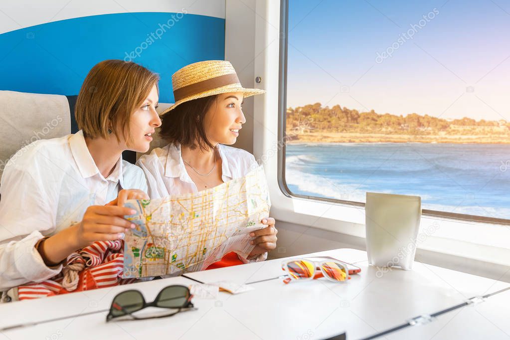 Two young girls of different race hold a map in their hands and look out the window of the train at the beautiful seascape. The concept of travel by the railroad on the sea coast