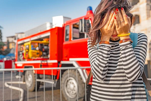 A woman in stress covers her face with her hands on the background of a fire fighting truck. Accident and insurance concept