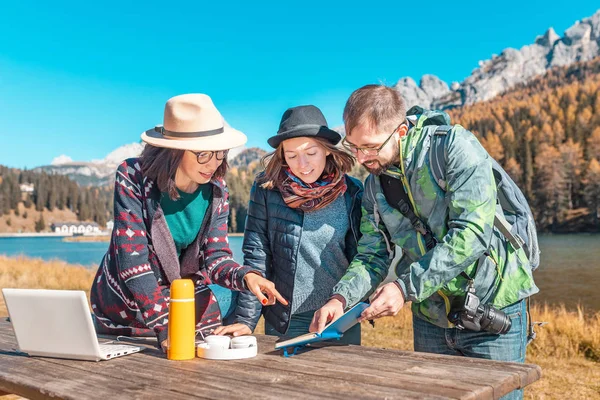 Three happy friends with notebook and laptop in nature park at the background of a mountain lake in Alps