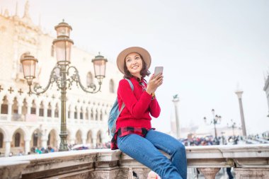Genç Asyalı kız mutlu traveler şapka ve Venedik'te San Marco meydanında parapetin üzerinde oturan sırt çantası. Tatil ve tatil İtalya ve Avrupa kavramı