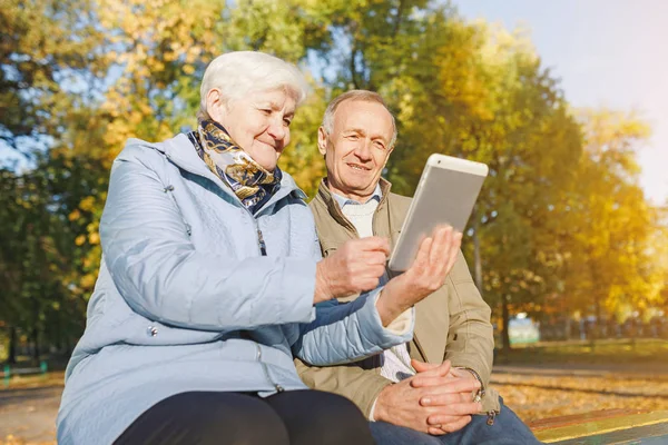 Senior couple sitting on a bench in autumn park and using digital tablet