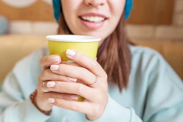 Une jeune fille dans un chapeau tricoté boit une boisson chaude dans une tasse en papier dans un café — Photo