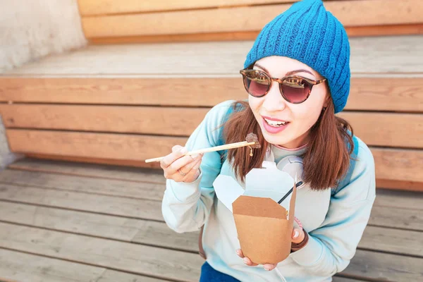 Модная азиатская девушка-хипстер ест восточную вок-лапшу палочками для еды. Концепция контейнеров для обеда и еды на вынос из дома из бумаги — стоковое фото