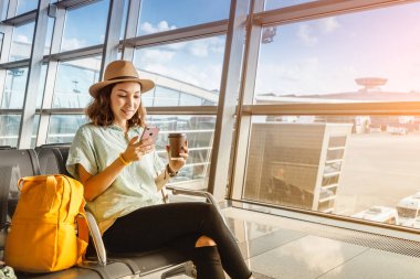 Asya kız tatil havaalanında ayrılış için bekliyor. Akıllı telefon ve içecek kahvesi kullanır