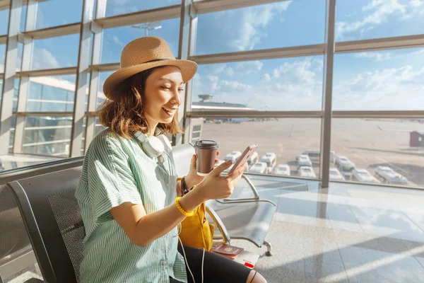 Asijská dívka čeká na odjezd na letišti na dovolené. Používá smartphone a pije kávu — Stock fotografie