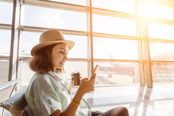 Азиатская девушка в ожидании вылета в аэропорту во время отпуска. Использует смартфон и пьет кофе — стоковое фото
