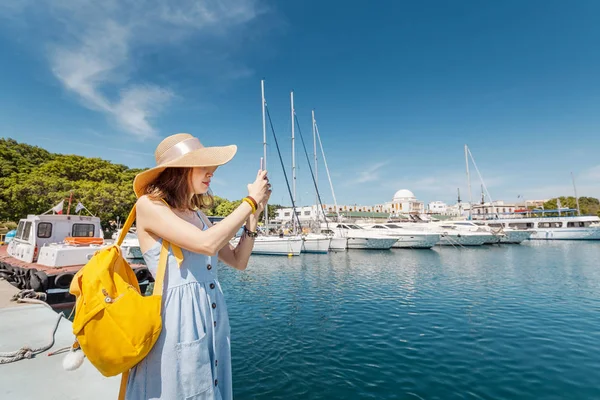 Young happy woman in hat with backpack standing near sea port with luxury yachts. Travel and vacation concept