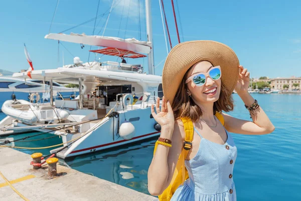 Joven mujer feliz en sombrero con mochila de pie cerca del puerto marítimo con yates de lujo. Concepto de viajes y vacaciones —  Fotos de Stock