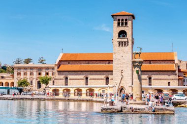 27 Mayıs 2019, Rodos, Yunanistan: Mandraki deniz limanına turist kalabalıkları ile Hirschkuh geyik heykeli ve kilise