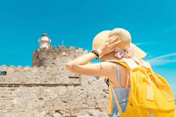 Mujer turista feliz de vacaciones posando con sombrero y mochila frente al antiguo fuerte y faro en ruinas en Rodas, Grecia — Foto de Stock