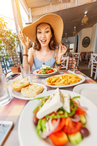 Glückliche asiatische Frau mit großem Hut beim Essen mit griechischem Salat horiatiki im Restaurant. Konzept der griechischen Küche — Stockfoto