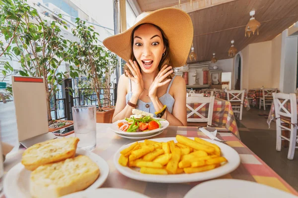 Счастливая азиатка в большой шляпе обедает с греческим салатом Хориатики в ресторане. Греческая кухня — стоковое фото