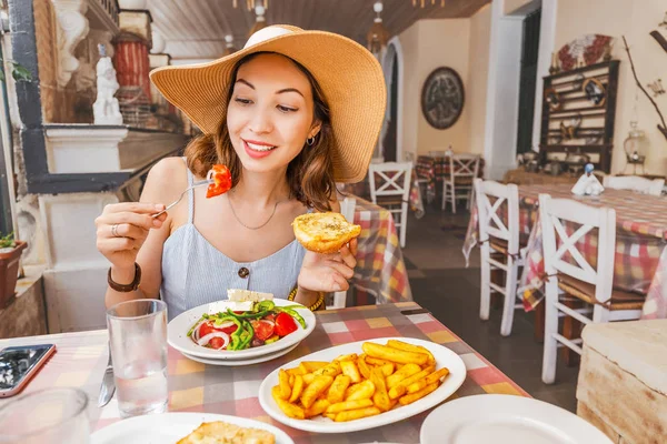 Happy asian woman in big hat having meal with greek salad Horiatiki in restaurant. Greece cuisine concept — Stock Photo, Image