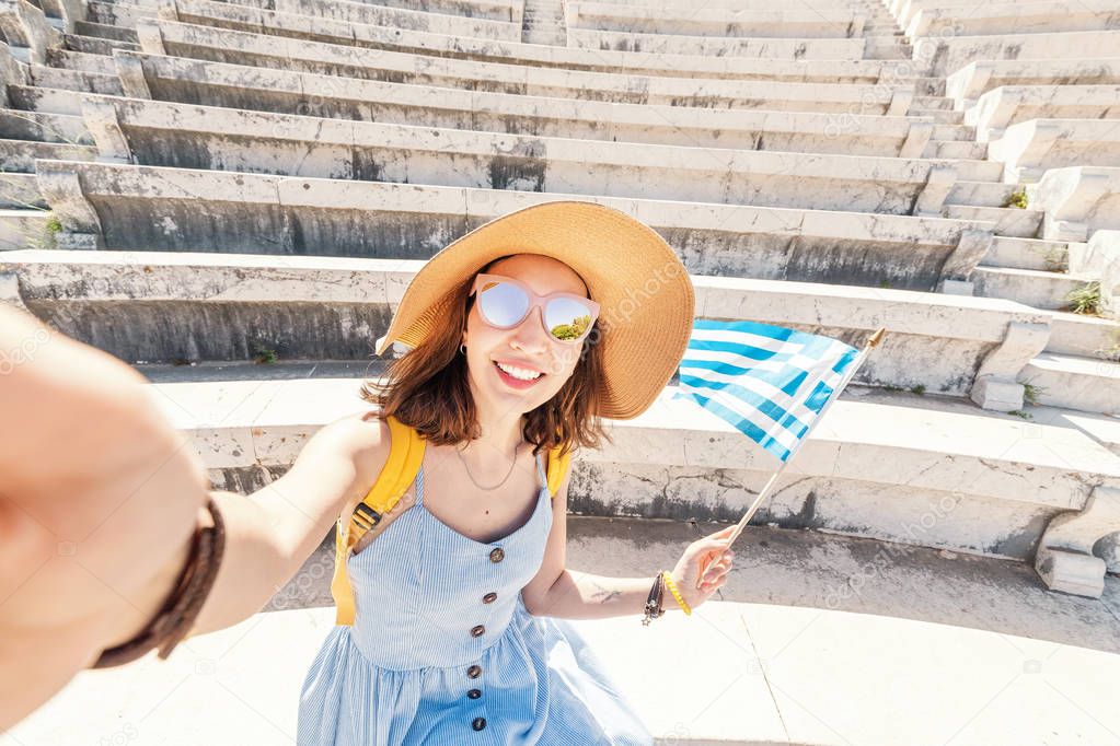 Asian girl tourist traveling and taking selfie photo in the ruins of an ancient Acropolis or the amphitheatre with a Greek flag. Active young travelers and students concept