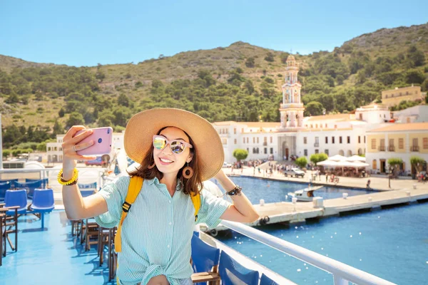 Young asian woman tourist making selfie photo with Panormitis monastery on the background from the cruise ship deck.