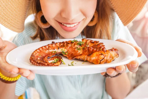 Happy asian woman in hat eating delicious grilled octopus in seafood restaurant. Delicacy and healthy gourmet cuisine — Stock Photo, Image