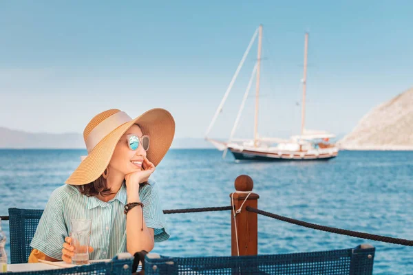 Lycklig kvinna klädd i stor hatt sitter i Café och ser till Seascape — Stockfoto