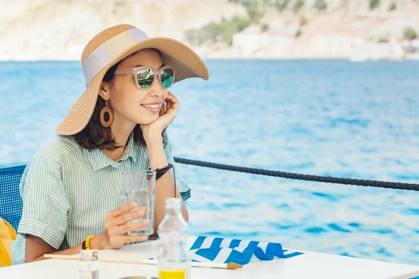 Glückliche Frau mit großem Hut sitzt im Café und blickt auf das Meer — Stockfoto