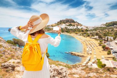 Genç ve mutlu bir kadın turist akıllı telefonuyla fotoğraf çekiyor kumlu bir sahil ve eski bir kasaba manzaralı. Tatil ve seyahat kavramı