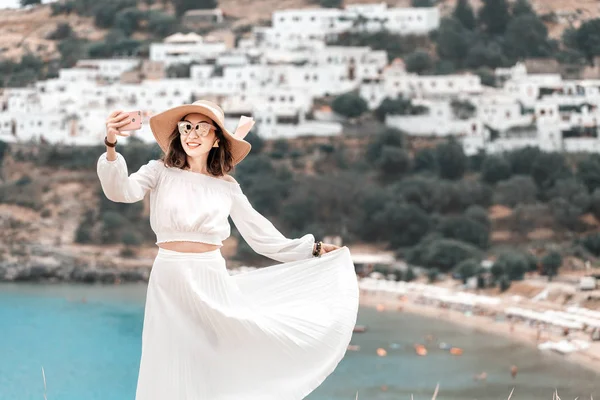 Elegantní turistická dívka, která si na pozadí starého bílého města Lindos v Řecku vzala fotografii na své smartphone — Stock fotografie