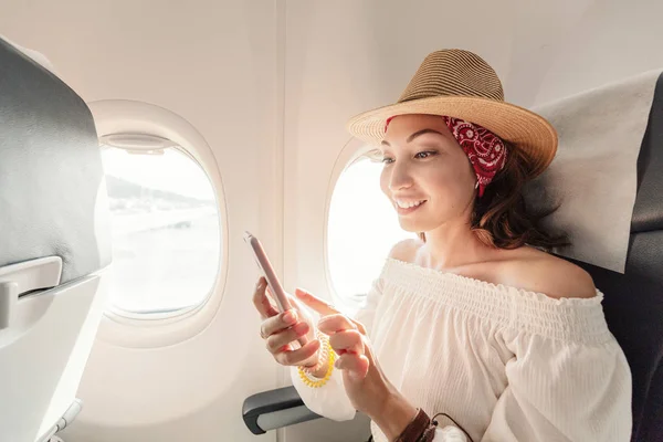 Asian woman passenger sitting in airplane near window and reading news from social networks or using travel applications in smartphone — Stock Photo, Image
