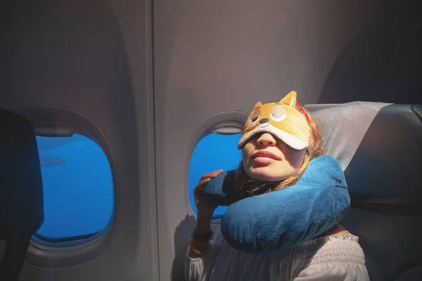 Glückliche und müde junge Frau, die bequem mit Maske und Kopfkissen im Flugzeug schläft. Ausgefeiltes Reisekonzept — Stockfoto