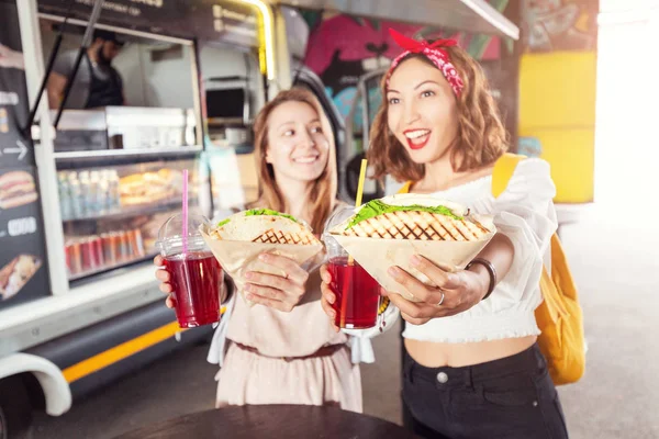 Two happy girls with appetizing fastfood snacks and drinks at the outdoor cafe — Stock Photo, Image