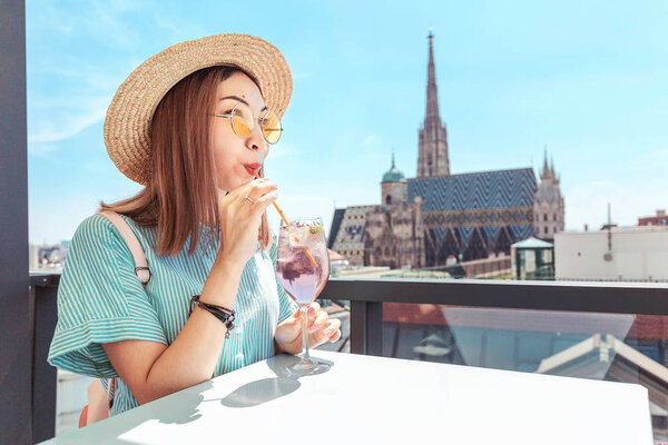 A cheerful girl in a hat drinks an alcoholic cocktail on the terrace of the bar with a great view of the city of Vienna with St. Stephen's Cathedral