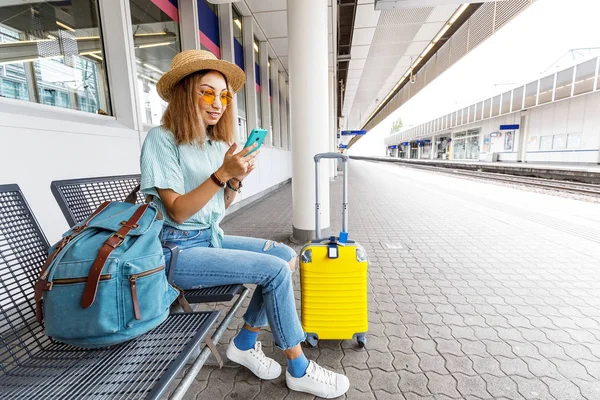 Cestovatel s asijskými dívkami se zavazadly sleduje jízdní řád vlaku a kupuje jízdenky na vlak pomocí elektronického použití telefonu při čekání na nástupiště stanice — Stock fotografie