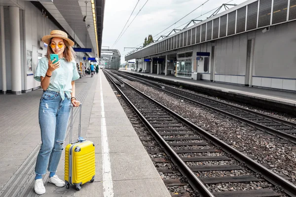 Cestovatel s asijskými dívkami se zavazadly sleduje jízdní řád vlaku a kupuje jízdenky na vlak pomocí elektronického použití telefonu při čekání na nástupiště stanice — Stock fotografie