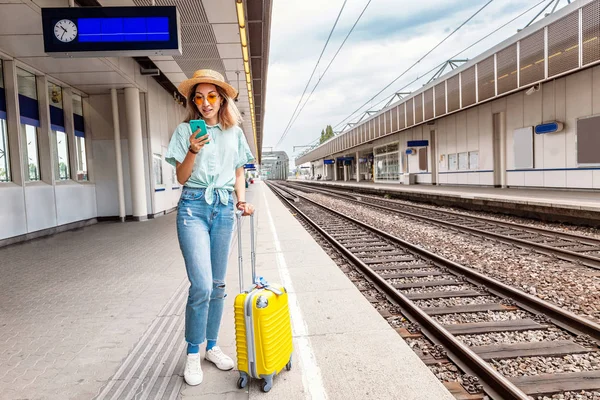 Cestovatel s asijskými dívkami se zavazadly sleduje jízdní řád vlaku a kupuje jízdenky na vlak pomocí elektronického použití telefonu při čekání na nástupiště stanice — Stock fotografie