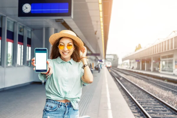 Ασιατικό κορίτσι ταξιδιώτη με αποσκευές παρακολουθεί το πρόγραμμα τρένων και αγοράζει εισιτήρια τρένων χρησιμοποιώντας την ηλεκτρονική εφαρμογή από το smartphone της, ενώ περιμένει στην πλατφόρμα του σταθμού — Φωτογραφία Αρχείου