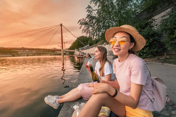 Dos amigas felices celebrando y brindando y bebiendo vino de unas pequeñas botellas y viendo un atardecer sobre el río Saone en la ciudad de Lyon, Francia — Foto de Stock