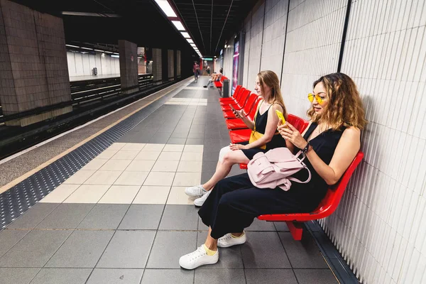 Duas amigas lendo smartphon no metrô enquanto esperam o trem. Conceito de vício em redes sociais e internet — Fotografia de Stock