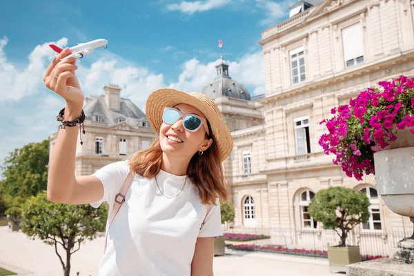 Ευτυχισμένος Ασιάτης/ισσα γυναίκα διαφημίζει Φτηνές πτήσεις ή αεροπορικές εταιρείες που παίζουν με ένα μικρό αεροπλάνο με φόντο το παλάτι του Λουξεμβούργου στο Παρίσι — Φωτογραφία Αρχείου