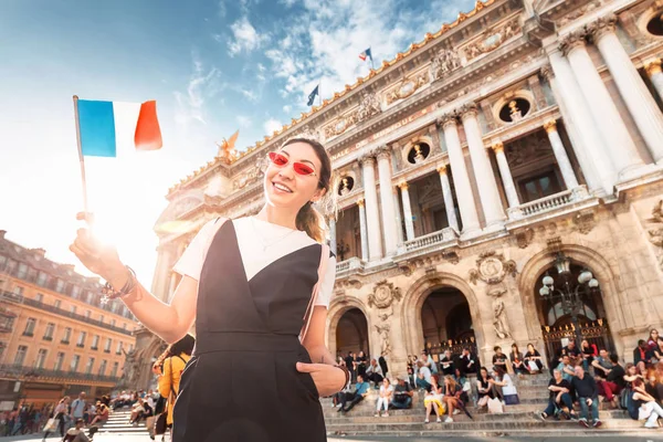 Menina asiática feliz viajante com bandeira francesa perto da Fachada principal da Ópera Garnier no edifício histórico da Academia de música de Paris. Desfocado pessoas irreconhecíveis em segundo plano — Fotografia de Stock