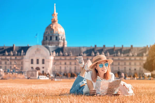 Asya mutlu kız gezgin bir çim üzerinde yatan ve Paris'te Invalides yakın harita okuma. Fransa'da yaşam tarzı ve turizm — Stok fotoğraf