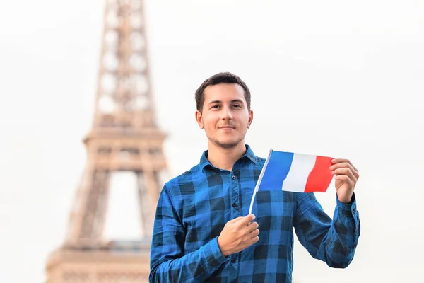 Leende ung man som innehar en fransk flagga på bakgrunden av den berömda Eiffeltornet. Resor, invandring och studier i Paris konceptet — Stockfoto