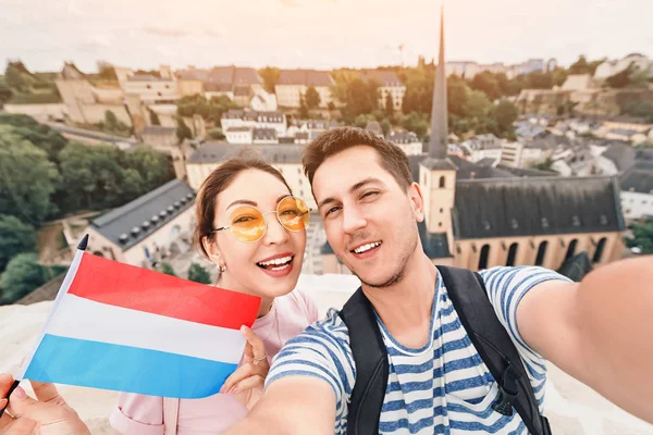 A happy couple of young people hug and take selfies with the flag of Luxembourg while sightseeing in old town. Tourism, immigration and education for students in Europe concept