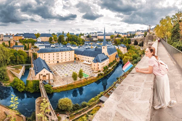 Uma menina viajante asiática feliz segura a bandeira de Luxemburgo e admira a grande vista panorâmica da cidade de Grund a partir do mirante. Turismo, recreação e vida no conceito de país — Fotografia de Stock