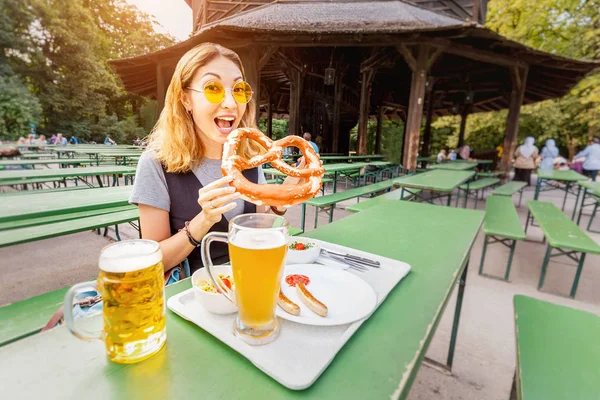 Asian girl eating traditional Pretzel and drinking fresh Bavarian beer in beer garden in Munich. The concept of traditional food festival and tourism