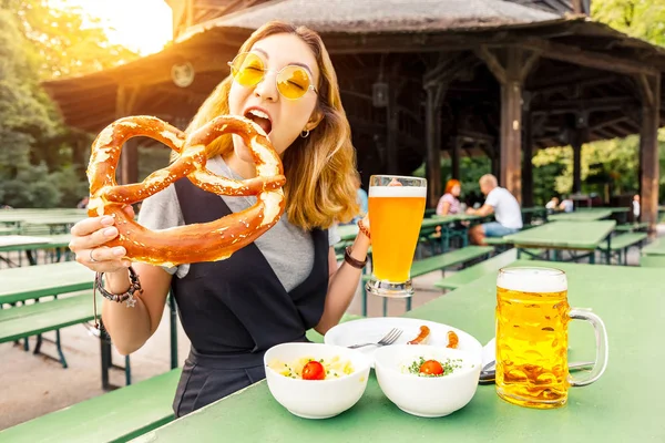 Asian girl eating traditional Pretzel and drinking fresh Bavarian beer in beer garden in Munich. The concept of traditional food festival and tourism