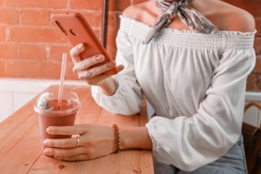 Mutlu Asyalı kız bir kafede smoothie içiyor ve akıllı telefonuyla sosyal ağlarda haberleri okuyor..