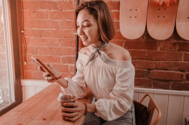 Mutlu Asyalı kız bir kafede smoothie içiyor ve akıllı telefonuyla sosyal ağlarda haberleri okuyor..