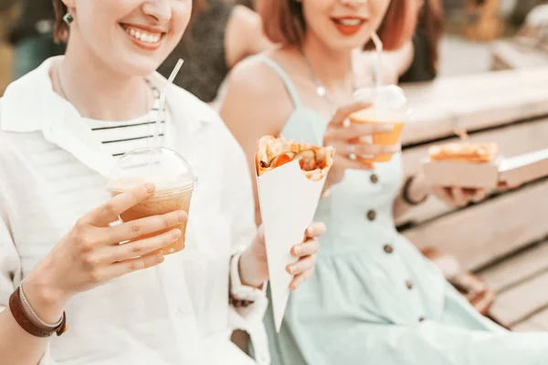 Joyful women friends relax in the Park and eat street food to take away and wash down with soft drinks. Casual communication and fast food holiday.