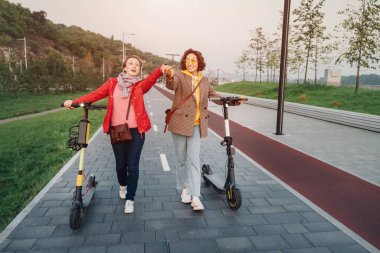 İki bayan arkadaş şehrin sokaklarında modern kiralık elektrikli scooterlara biniyor. Ulaşım ve yaşam tarzı ve arkadaşlık kavramı