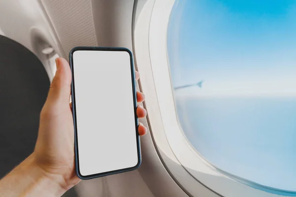 A man's hand holds a smartphone with a cut-out white blank screen against the background of an airplane window and blue sky during a vacation flight. Concept of communication and travel apps