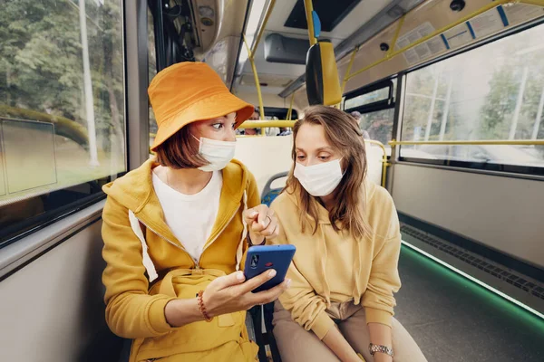 Two female friends in medical masks travel on public transport during the coronavirus outbreak and surf the Internet via smartphone